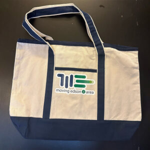 A beige and navy blue tote bag with long handles. The front features a logo with green and blue lines and the text moving edson area printed below the logo. The bag is placed on a dark surface.