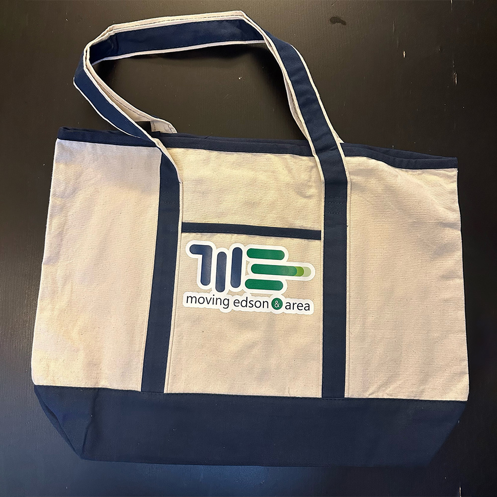 A beige and navy blue tote bag with long handles. The front features a logo with green and blue lines and the text moving edson area printed below the logo. The bag is placed on a dark surface.